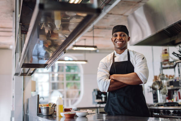 Chef with his arms crossed standing at restaurant kitchen. Male cook wearing apron standing by kitchen counter and smiling.